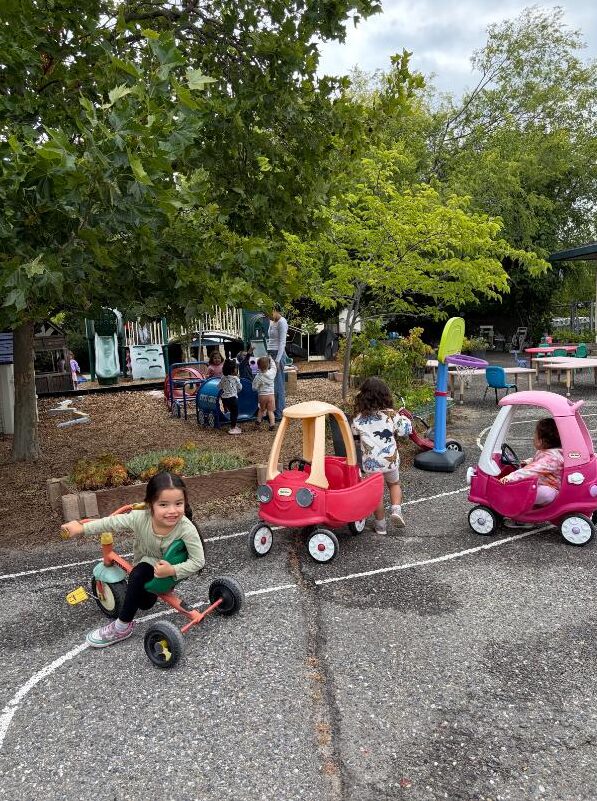 Children riding trikes and ride-on cars in the play yard